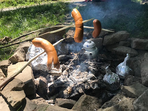 Cervelats cooking on sticks over an open fire, pot of tea and roasted peaches in the embers