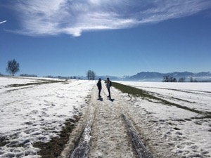 Two figures in silhouette on a snowy path under blue skies, with mountains in the background.