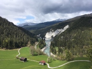 View down the Vorderrhein valley. Green pastures, next to river with crumbling cliffs in background. 