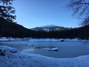 Sun setting over distant mountains. Frozen lake in foreground framed by trees.