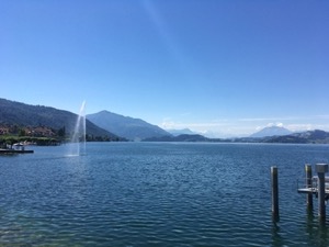 Fountain and blue waters of the Zuggersee, under blue skies, Rigi and Pilatus mountains in the background