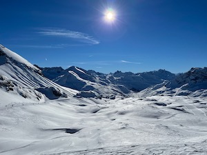 Snow covered mountains under bright blue skies. Ski piste visible snaking down the valley.