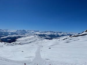 View down a ski run. Mountains in the background, blue skies.