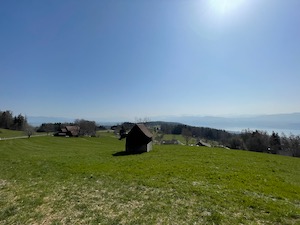 Swiss hut in a green field, under blue skies. Alps barely visible in the background through haze.