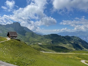 Alpine hut sitting above green meadows, mountains in the background sunny skies.